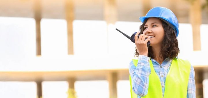 A woman wearing a light blue plaid shirt, yellow safety vest, and light blue hard hat smiles while holding up a radio.