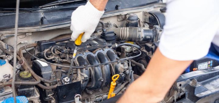 An auto mechanic wearing white gloves, using a flat-head screwdriver to perform repairs to a car’s engine.