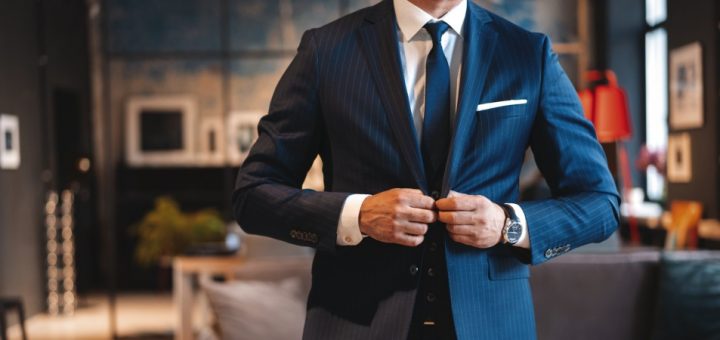 Man buttoning his blue suit jacket and wearing a tie, a white shirt, and a watch inside a nice room with a couch.