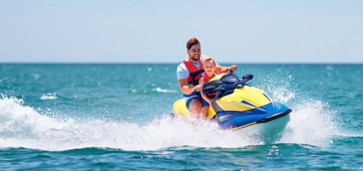 A father and sun riding a jet ski on the water on a bright and sunny day. The water is splashing around their jet ski.