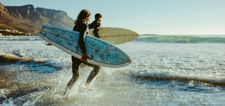 A man and woman running toward the ocean wearing neoprene wet suits carrying surfboards under their right arms.