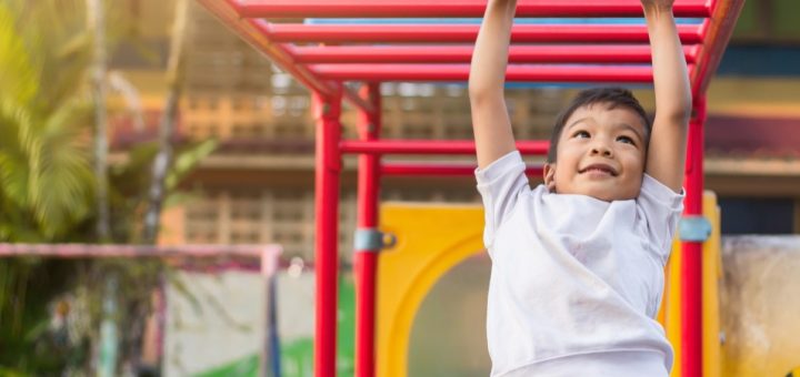 A young, school-aged boy is hanging from red monkey bars on the playground. The rest of the playground is blurry.