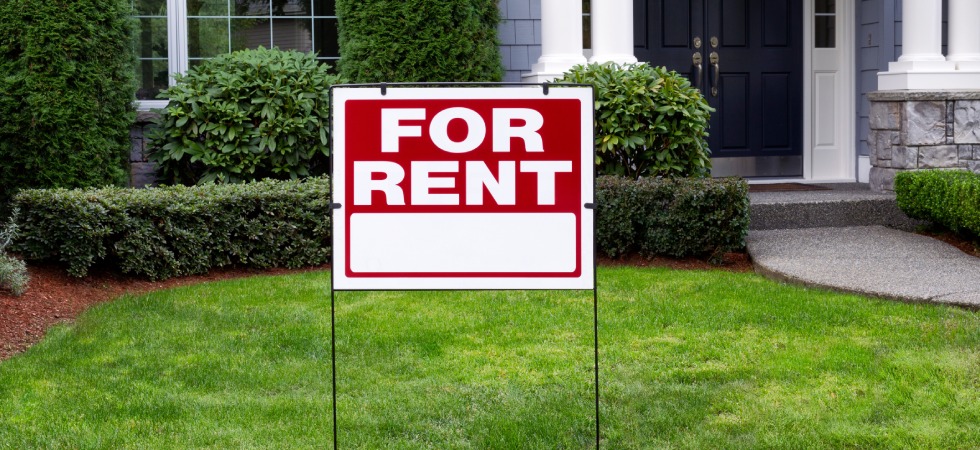A sign that reads for rent sits in the front yard of a home. Meticulously trimmed bushes appear in the background.