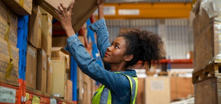 A young woman is placing a cardboard box on a warehouse shelf. She is wearing a blue shirt and a bright yellow vest.