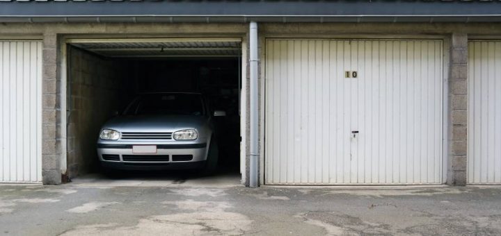 A silver car sits inside a concrete storage unit with the garage door open. The storage facility has a black roof.
