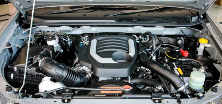 A silver car has its hood open and propped up. There are many components, and the large engine sits in the middle.