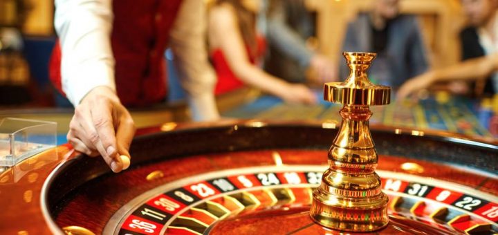 A close-up of a croupier's hand holding a small ball above a roulette wheel. There are people at another table in the background.