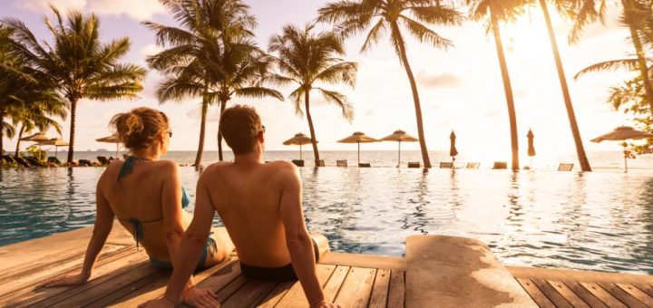 A couple sitting at the edge of a deck over the water with the ocean and palm trees in the background as the sun sets.