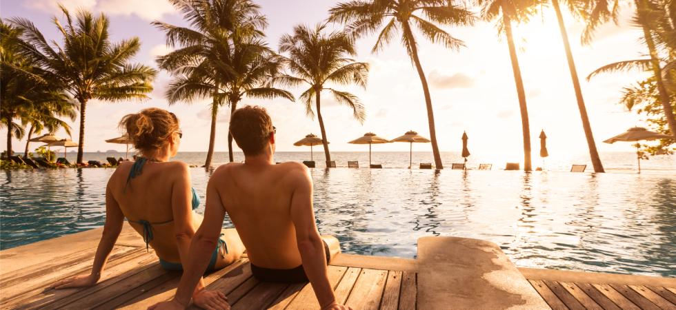 A couple sitting at the edge of a deck over the water with the ocean and palm trees in the background as the sun sets.