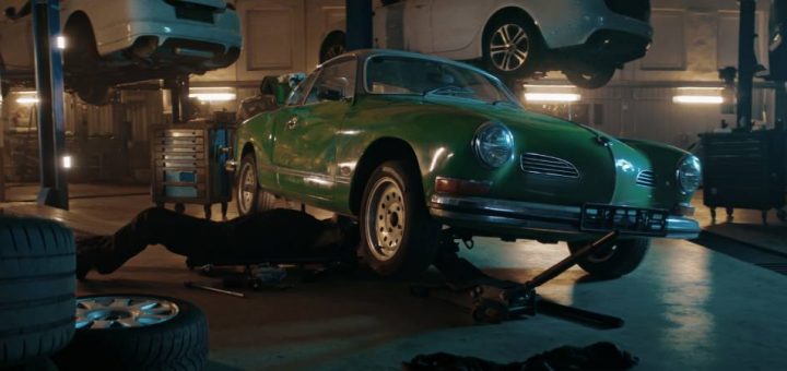 A person working under a green vintage car on a jack in a dimly lit workshop with modern cars in the background.