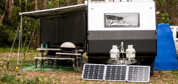 A parked RV camper in a forest where three solar panels are placed next to the vehicle. There is a grill and table nearby.