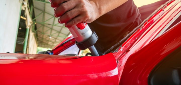 An auto body mechanic applying an adhesive to the windshield of a red car. They're using a caulk gun to apply the glue.