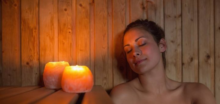 Woman wearing a white towel around body and head sitting inside a wooden sauna with two small candles.