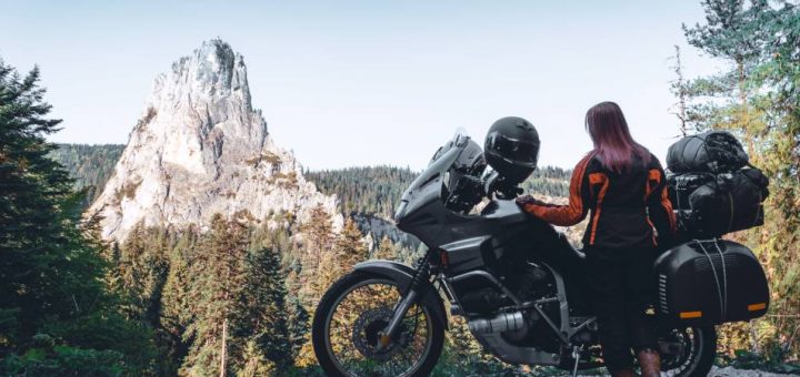 A young woman wearing an orange and black jacket stands behind a large motorcycle while admiring a rocky mountain.