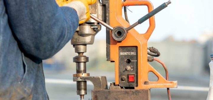 An individual wearing yellow gloves is operating a vertical drilling machine to cut a piece of steel.