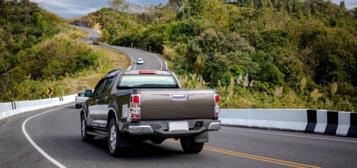 A pickup truck is seen from behind driving down a twisty mountain highway with other cars and lots of greenery.