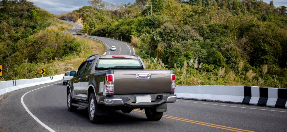 A pickup truck is seen from behind driving down a twisty mountain highway with other cars and lots of greenery.