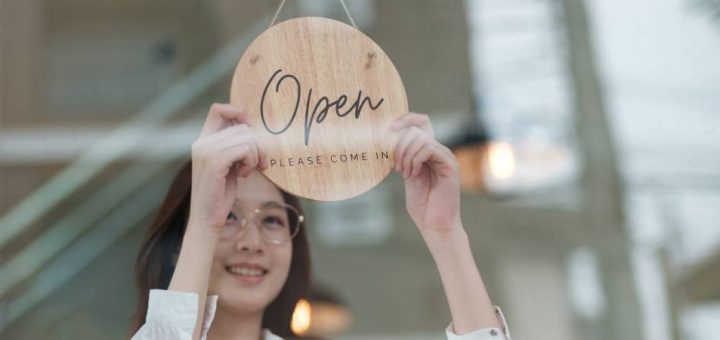 A woman flips a round wooden sign that reads "Open, Please Come In" in a shop window to indicate the business is open.
