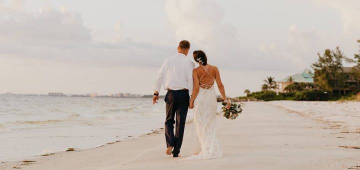 A newlywed bride and groom holding hands and walking on the beach, celebrating their wedding day at sunset.