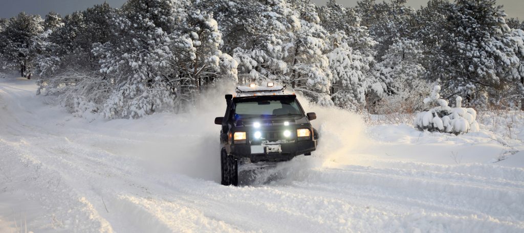 A black off-roading vehicle with bright hood lights and a roof rack drives through heavy snow piles in a forested area.
