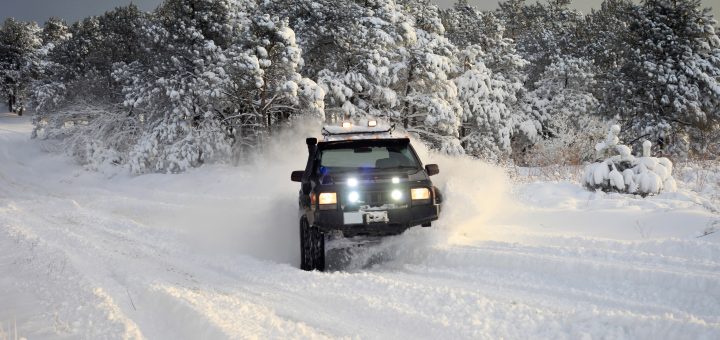 A black off-roading vehicle with bright hood lights and a roof rack drives through heavy snow piles in a forested area.