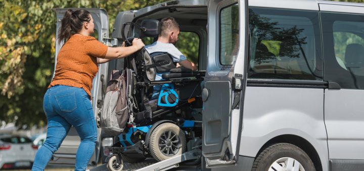 A woman in an orange shirt and jeans pushes a man in a wheelchair up a steel ramp into the back of a silver van.