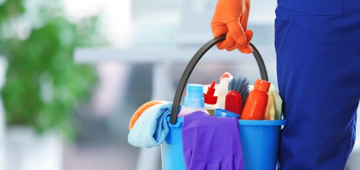Close-up of a hand wearing an orange latex cleaning glove holding a bucket full of plastic bottles, sponges, and cloths.