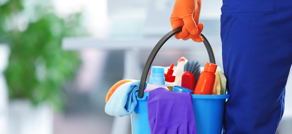 Close-up of a hand wearing an orange latex cleaning glove holding a bucket full of plastic bottles, sponges, and cloths.