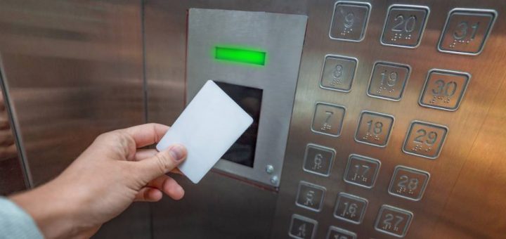 A person holds a blank, white key card up to an access control reader inside an elevator. A green light is above the reader.