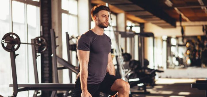 A man wearing black shorts and a gray T-shirt is holding two kettlebells while doing lunges in a gym.