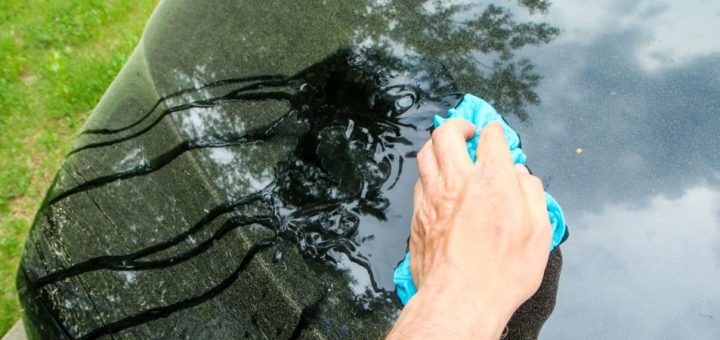 A person wiping down the hood of their car with a blue rag. The surface is covered with a layer of pollen.