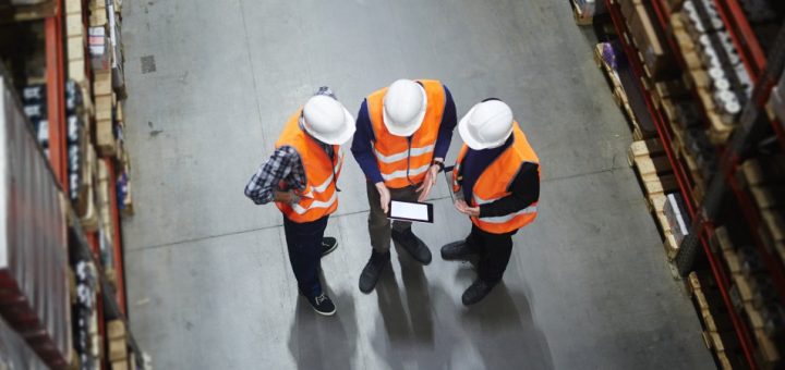 Aerial view of three warehouse workers in orange visibility vests looking at a tablet in the aisle between two storage racks.