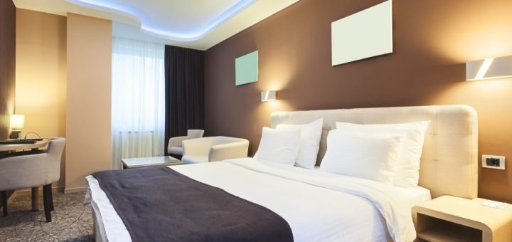 A well-lit hotel room features a bed with white pillows and sheets, as well as three tan chairs and a table.