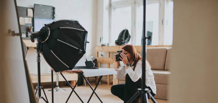 A woman crouches to take a picture of something on a table surrounded by professional photography equipment.