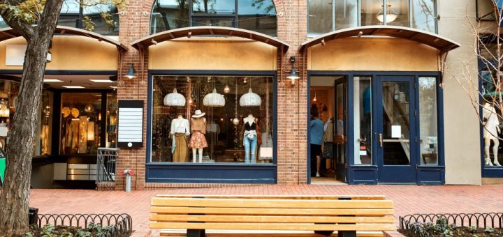 The exterior of a clothing shop with mannequins in the window. A wooden bench sits on the brick sidewalk out front.