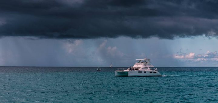 A boat in the middle of open waters moves along as a large, dark cloud looms over it. Low-hanging clouds are in the background.
