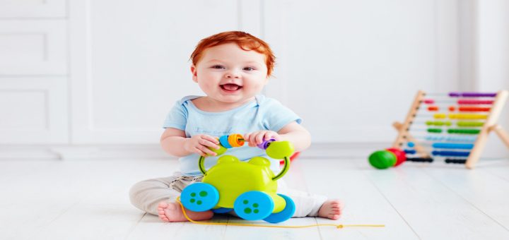A red-headed baby in a blue onesie smiling while playing with a toy and sitting among other toys in a white room.
