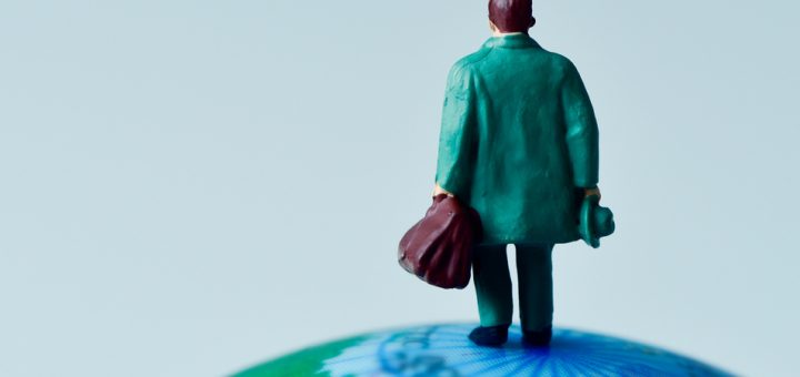 A small figurine of a man in business attire sits on top of a plastic globe. The figurine is visible from the rear.