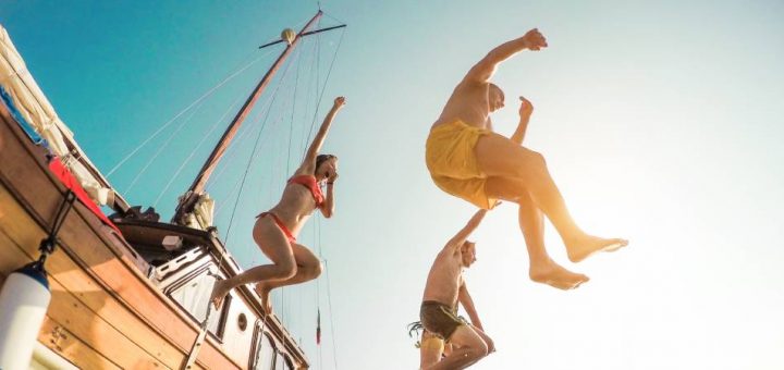 A group of people jump off the side of a small boat and into the water wearing their swimsuits.