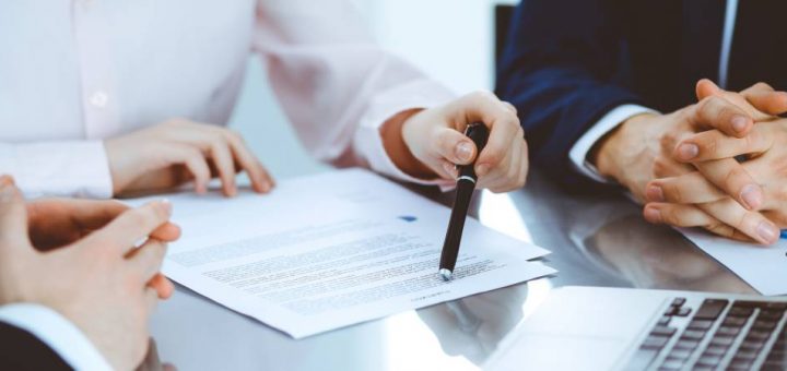 A group of people in suits sit at a table and negotiate the contract in front of them. One points at it with a pen.