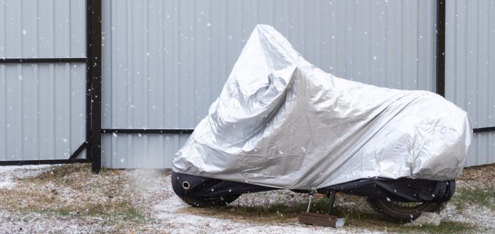 A motorcycle under a silver tarp sits in a fenced yard. Snow falls around the motorcycle and lightly covers the ground.