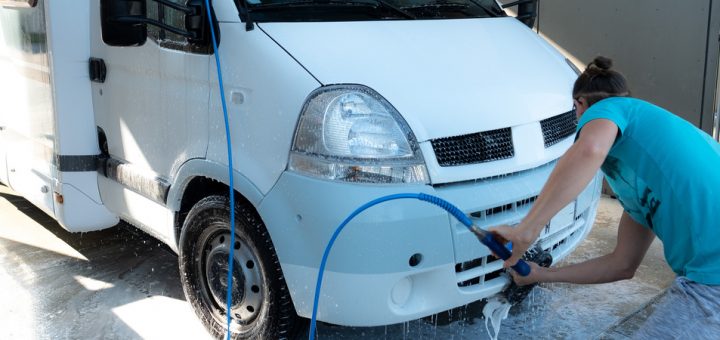 A man crouches down and uses a hose to rinse off the front of his RV. The RV mostly white and black.