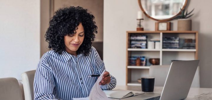 A woman reviewing some paperwork while sitting at a table. Her laptop, smart phone, and coffee cup are nearby.