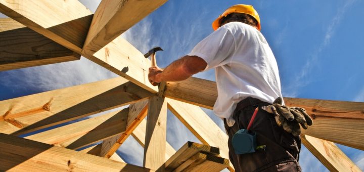 A man wearing a white T-shirt and an orange hard hat using a hammer to build a wooden structure on a sunny day.