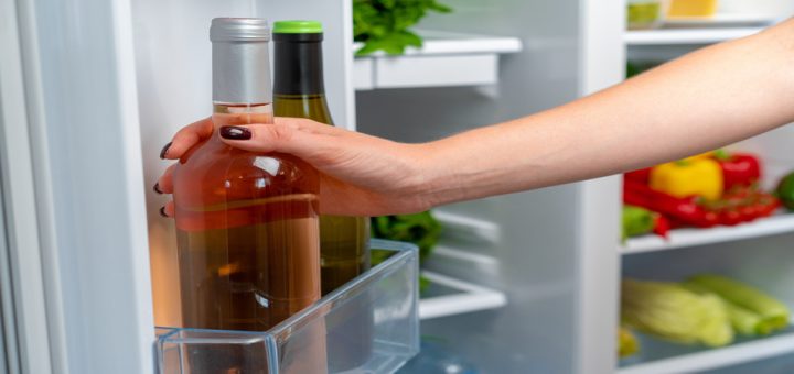 A hand with manicured nails reaches to pull a bottle of wine with no label on it out of a fridge.