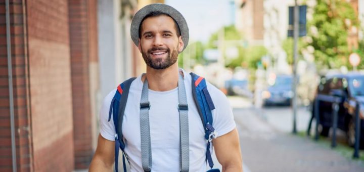 A male tourist walks the streets of a town while wearing a backpack and a camera around his neck. He holds a phone.