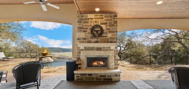 A large stone patio with an outdoor fireplace, chairs, and a ceiling fan near a backyard with a pool.