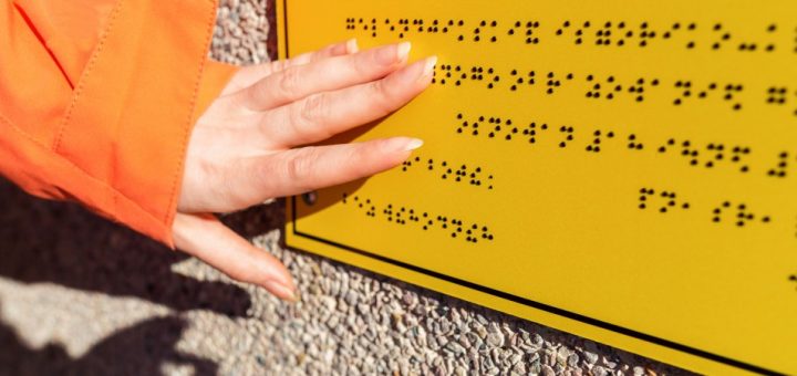 An individual in an orange jacket using their fingers to read the black braille characters on a yellow sign.