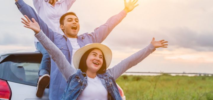 Three friends stand with their arms raised in celebration while standing outside their car during a road trip stop.