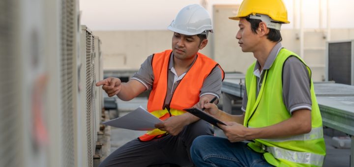 Two engineers with a clipboard and papers check equipment on the top of a roof. They wear hard hats and safety vests.